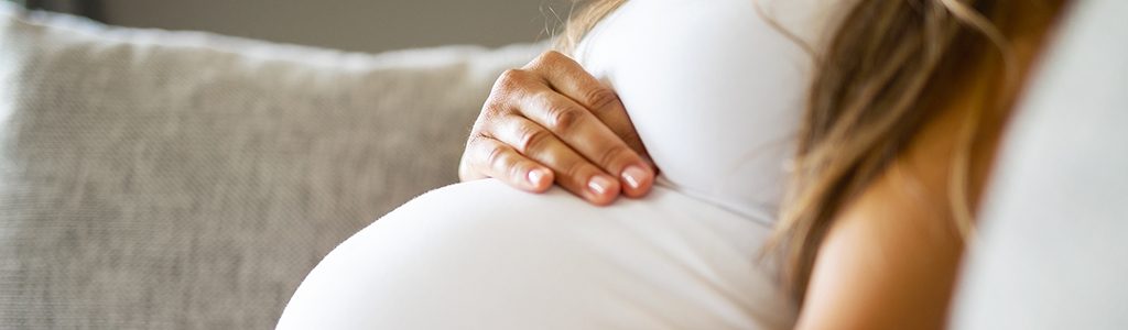 Close-up of pregnant woman relaxing and sitting on the side on the sofa. Holding a hands on the tummy.